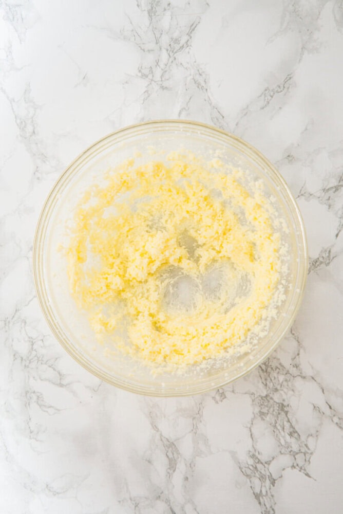 A glass bowl containing a mixture of creamed butter and sugar sits on a white marble surface.
