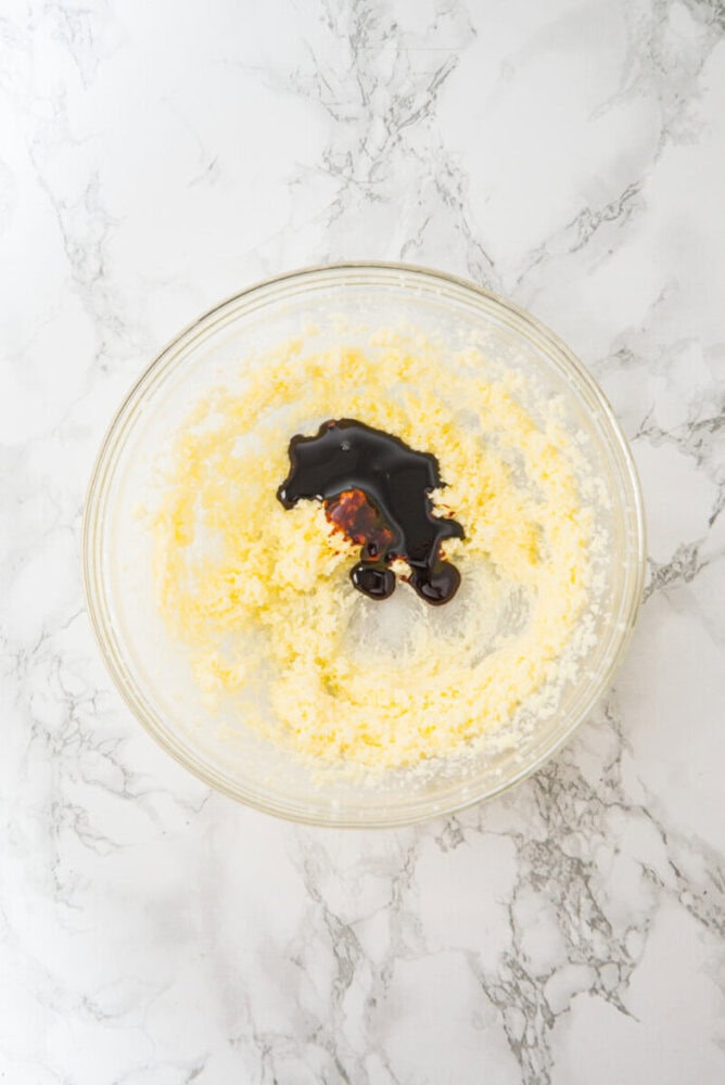 A glass bowl on a marble surface containing creamed butter and sugar with a splash of dark liquid, likely vanilla extract, on top.