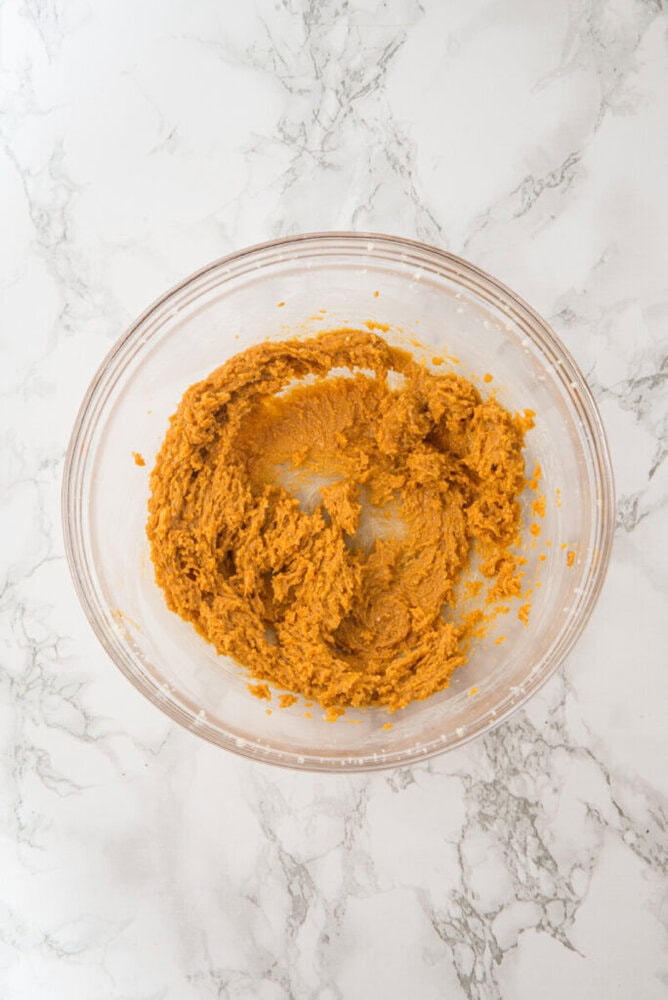 A glass bowl with light brown cookie dough mixture sits on a white marble surface.