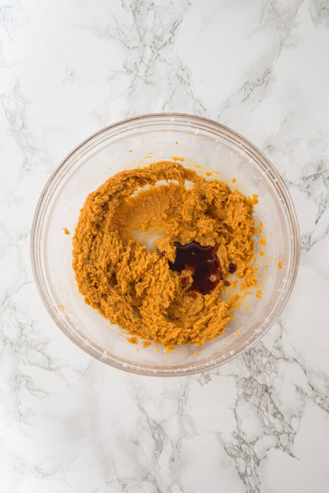 A glass bowl containing partially mixed brown batter with a small pool of liquid vanilla extract on top, placed on a white marble surface.
