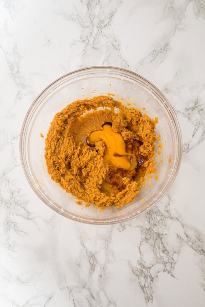 A glass bowl containing brown cookie dough mixture with egg yolk cracked on top, placed on a white marble surface.