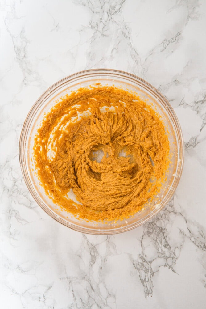 A glass bowl with creamed brown cookie dough mixture sits on a white marble surface; two circular indentations and a curved line form a smiley face in the dough.