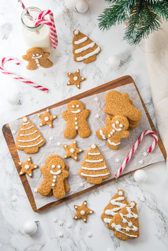 Assorted gingerbread cookies shaped like people, trees, and stars with white icing, arranged on a wooden board with candy canes and a glass of milk.