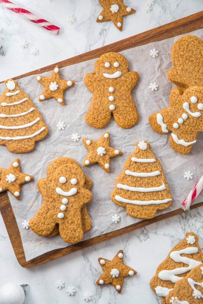 A wooden tray with iced gingerbread cookies shaped like trees, stars, and gingerbread people, on parchment paper, with candy canes and more cookies around.