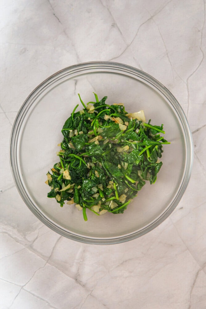 A glass bowl containing saut&eacute;ed spinach mixed with chopped onions, set on a light-colored marble surface.