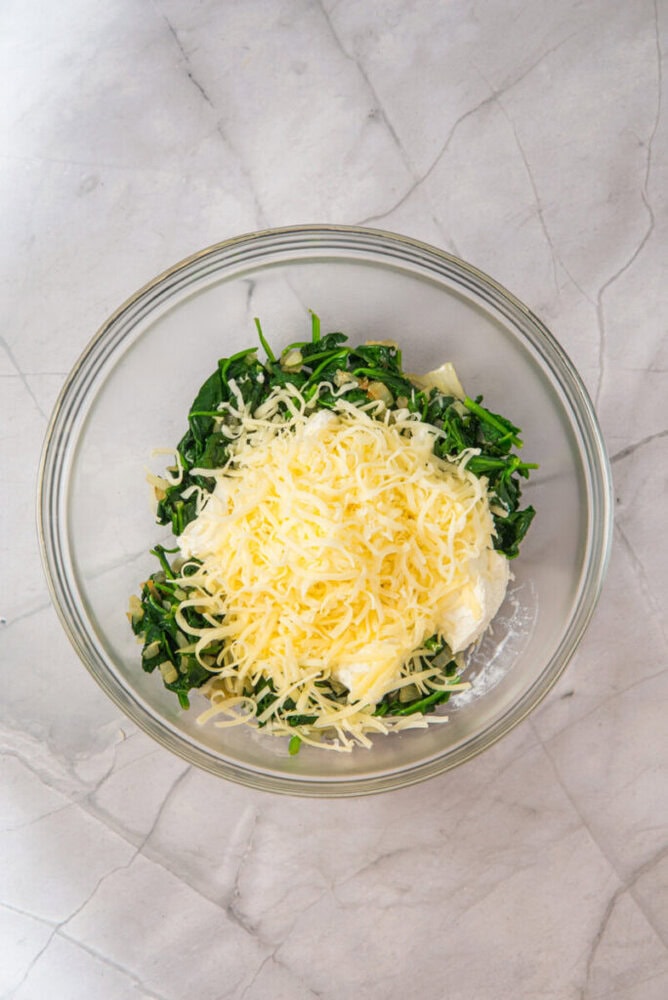 A glass bowl containing chopped spinach, shredded cheese, and cream cheese sits on a light-colored marble countertop.