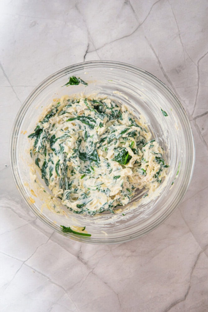 A glass bowl containing a creamy mixture with visible green spinach leaves, placed on a light-colored marble surface.