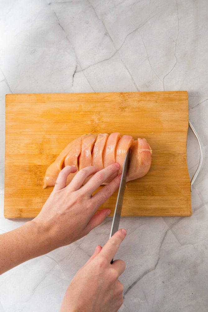 A hand slices a raw chicken breast into strips on a wooden cutting board with a knife, on a marble countertop.