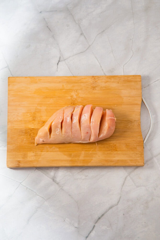 A raw chicken breast with several diagonal cuts is placed on a wooden cutting board on a light marble surface.