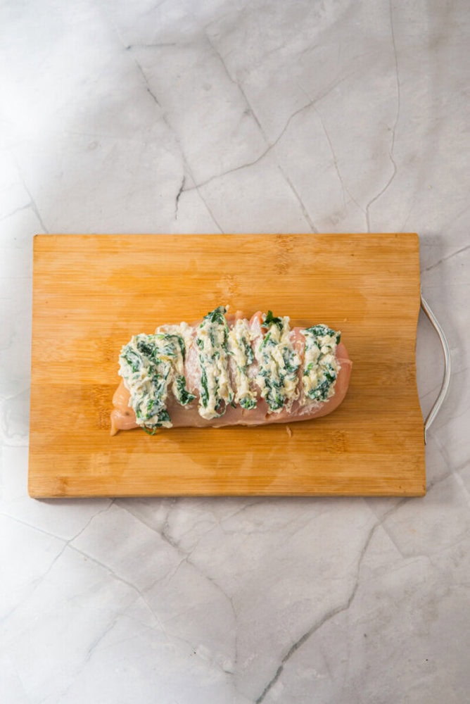 A raw chicken breast with slits is filled with a spinach and cheese mixture, placed on a wooden cutting board on a marble countertop.