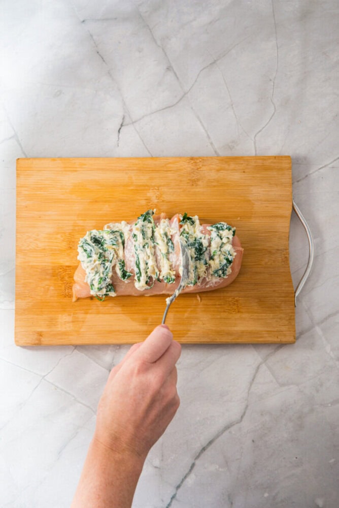 A hand spreads a spinach and cheese mixture onto a butterflied chicken breast on a wooden cutting board.