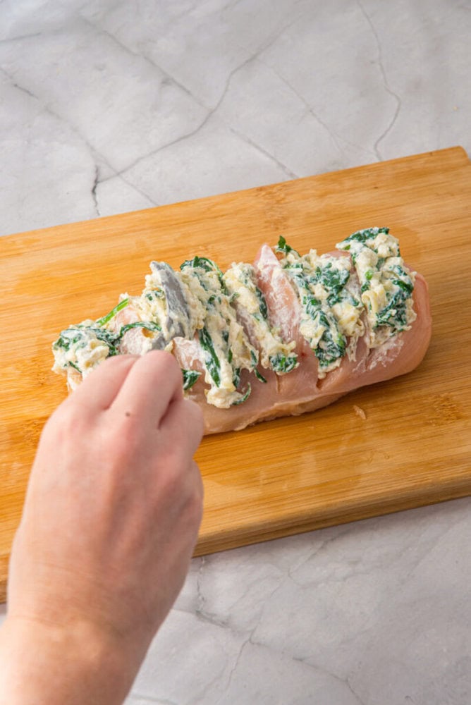 A hand spreads a spinach and cheese mixture into slits cut in a raw chicken breast on a wooden cutting board.