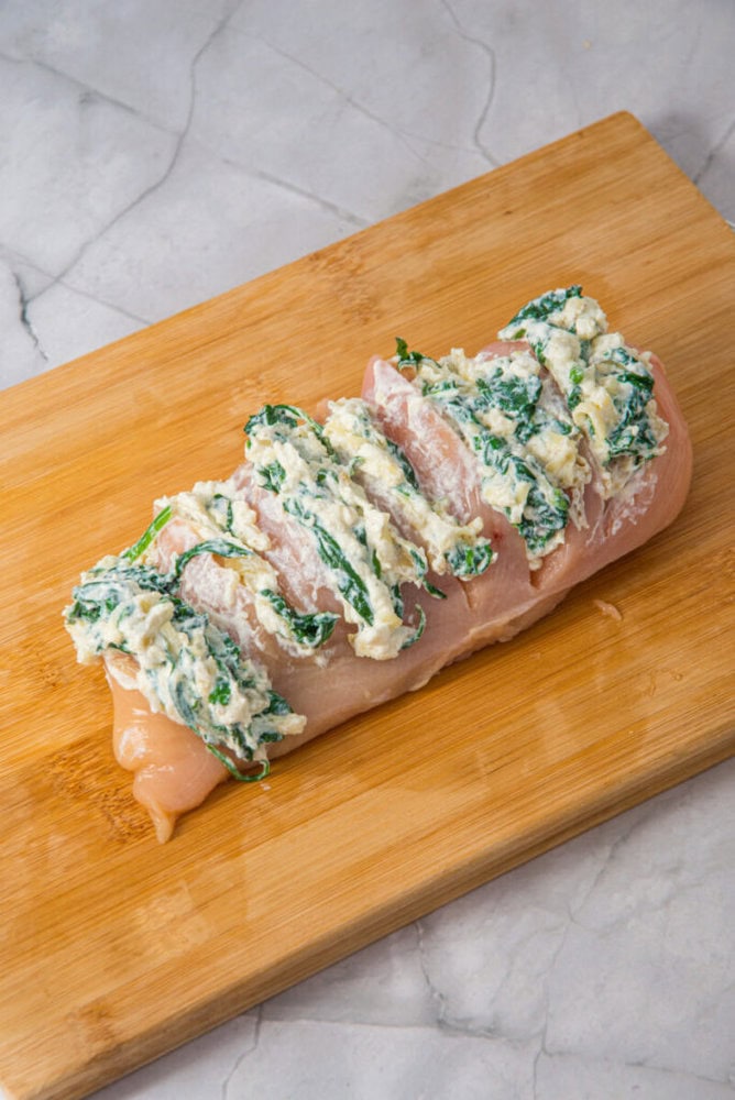 Raw chicken breast sliced and stuffed with a spinach and cheese mixture on a wooden cutting board, placed on a marble countertop.
