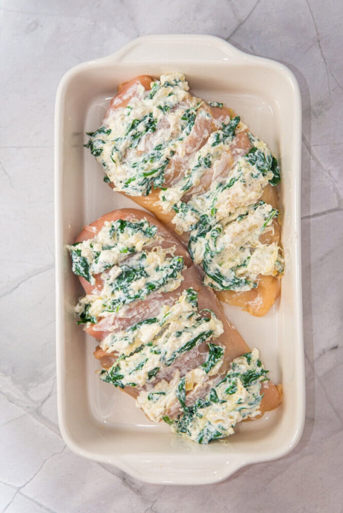 Two raw chicken breasts in a white baking dish, each topped with a mixture of spinach and creamy cheese filling.