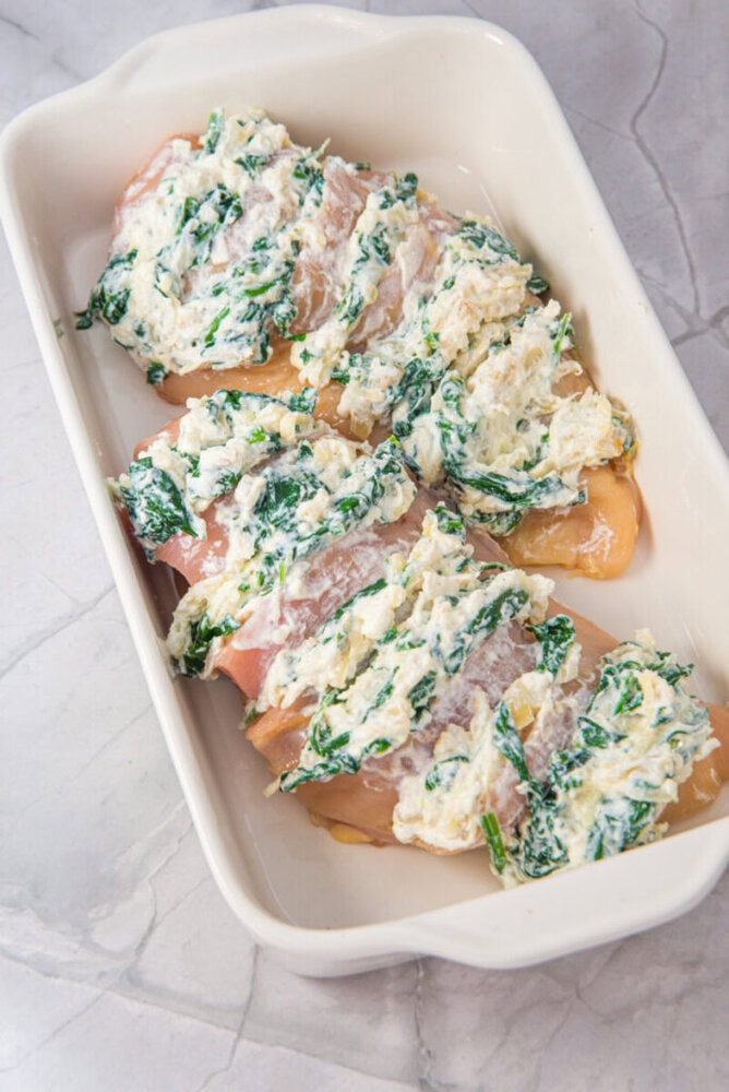 Three raw chicken breasts in a white baking dish, each topped with a spinach and cheese mixture, prepared for baking.
