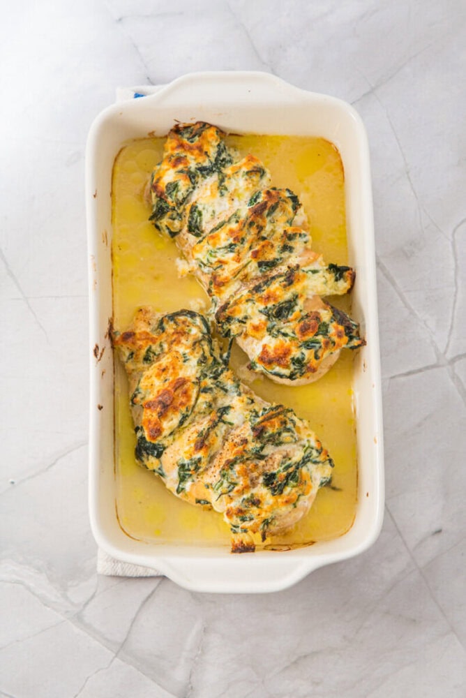 Three baked chicken breasts topped with a creamy spinach mixture in a white rectangular baking dish on a light marble surface.