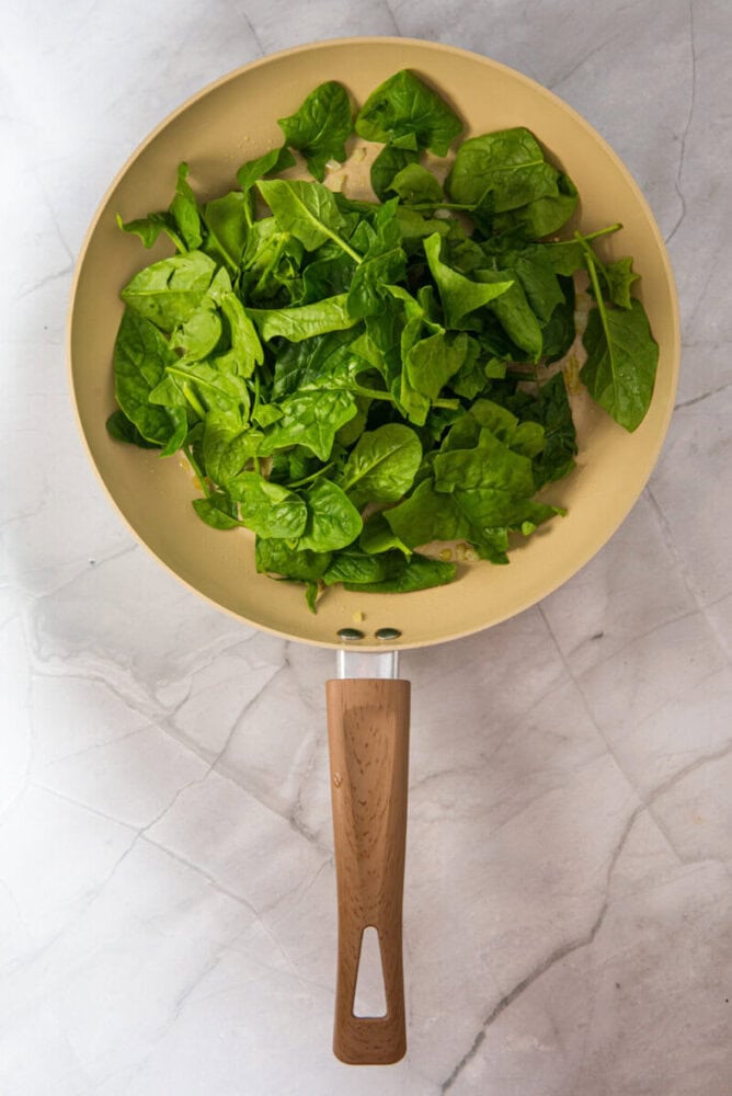A beige frying pan with fresh spinach leaves sits on a marble countertop, viewed from above.