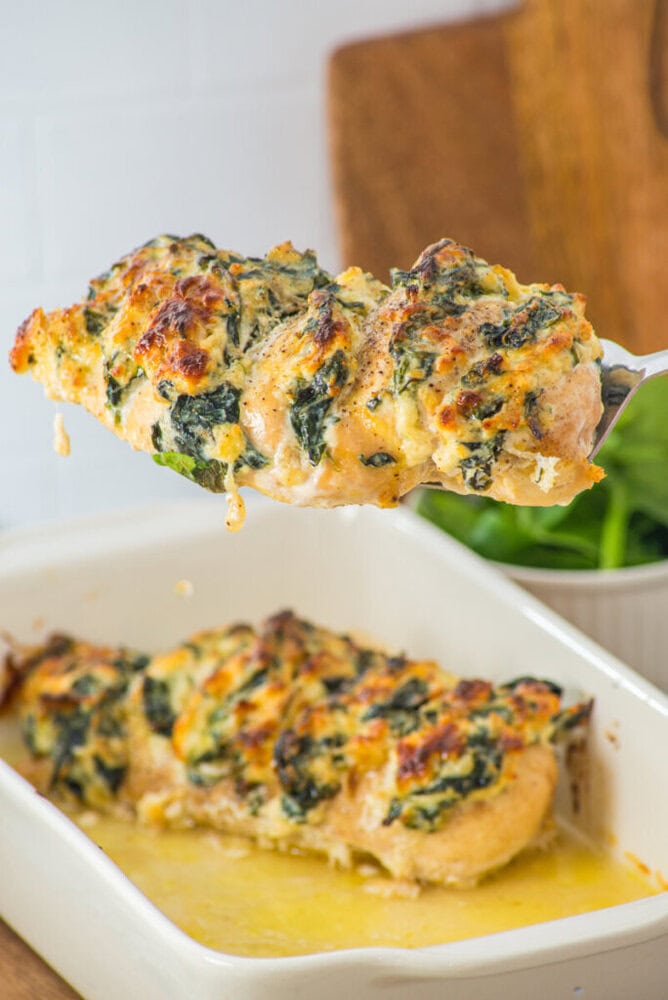 A close-up of a stuffed chicken breast with melted cheese and spinach being lifted from a baking dish, with another stuffed chicken breast in the dish below.