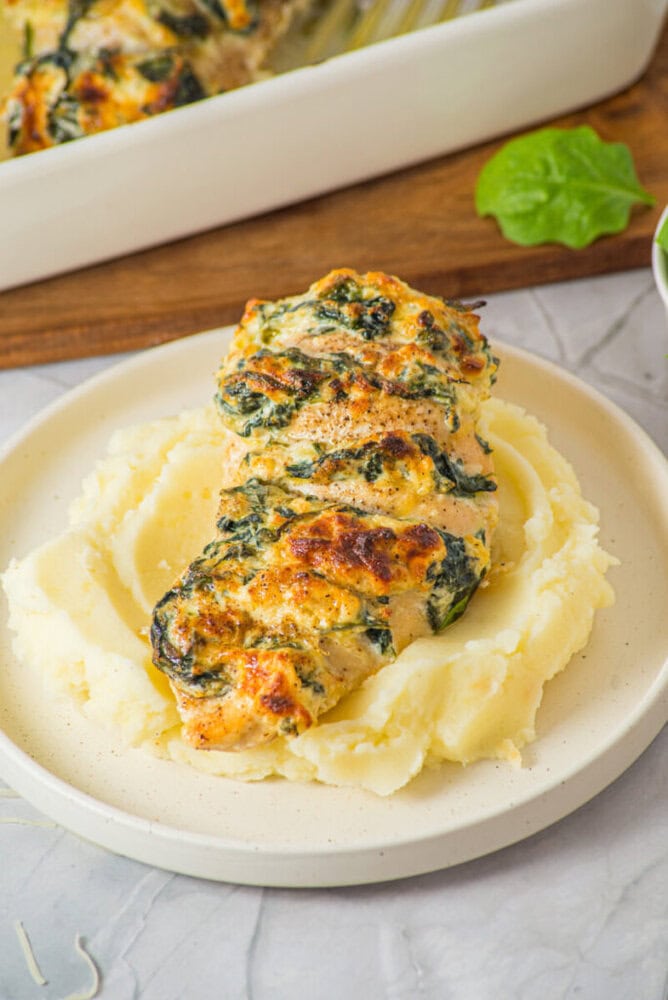 A piece of baked spinach and cheese stuffed chicken breast served on a bed of mashed potatoes on a white plate.