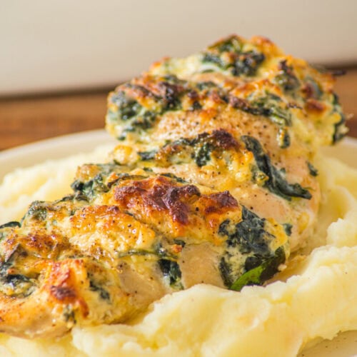 A stuffed chicken breast with cheese and spinach sits on a bed of mashed potatoes on a beige plate.