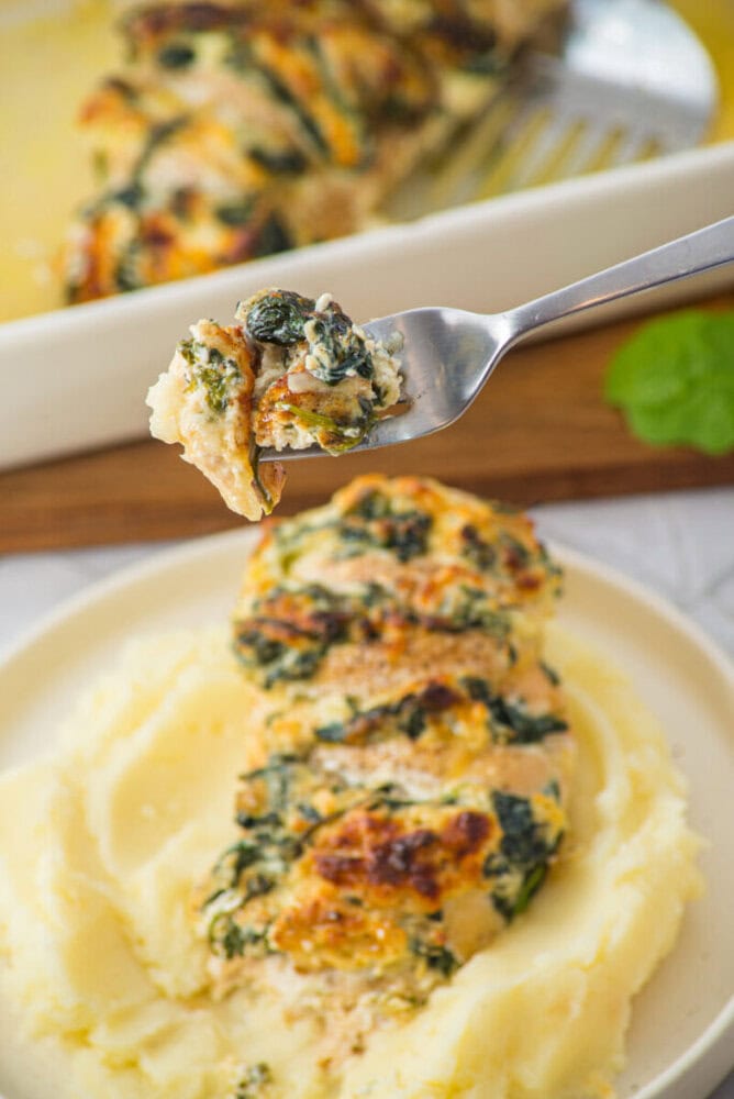 A fork holds a piece of chicken stuffed with spinach and cheese above a plate of mashed potatoes; a baking dish is in the background.