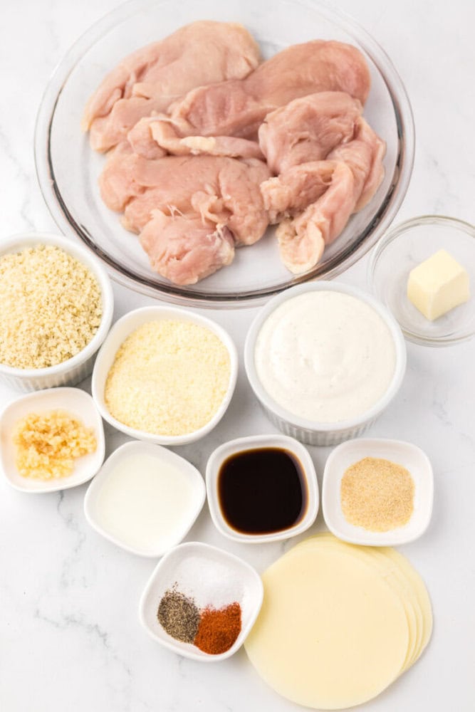 Raw chicken breasts in a glass bowl with small dishes of panko breadcrumbs, grated cheese, garlic, cream, Worcestershire sauce, butter, milk, spices, and sliced cheese on a white surface.