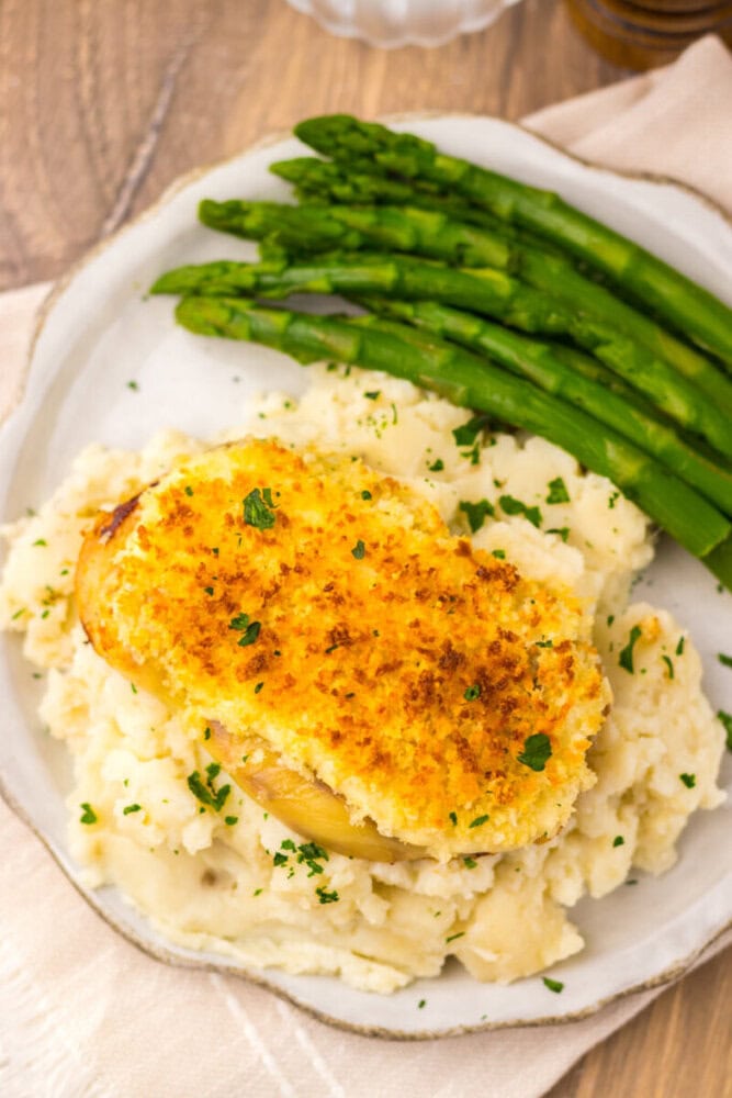 A plate of breaded, baked chicken breast on mashed potatoes, served with steamed asparagus spears on the side.