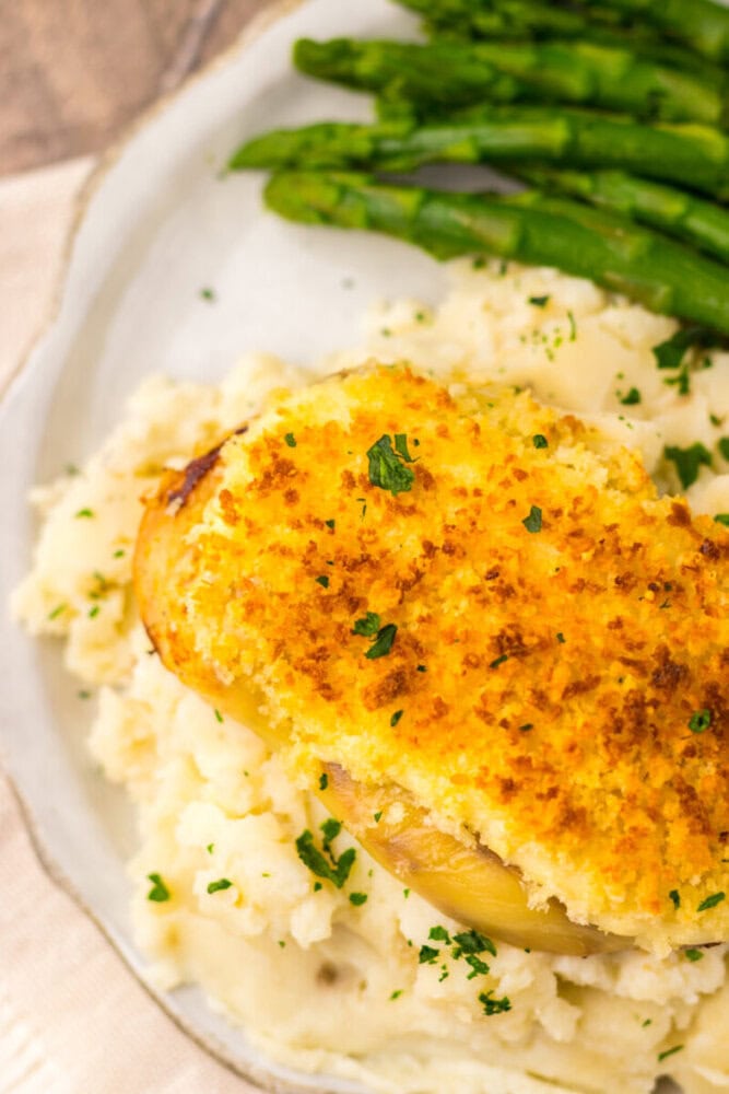 A plate of mashed potatoes topped with a breaded, baked chicken breast, garnished with parsley, and served with steamed asparagus.