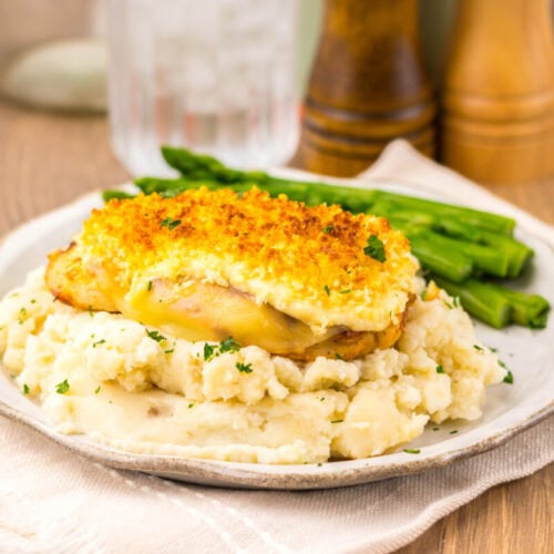 A plate with baked chicken topped with bread crumbs on mashed potatoes, served with asparagus spears. Salt and pepper shakers and a glass of water in the background.