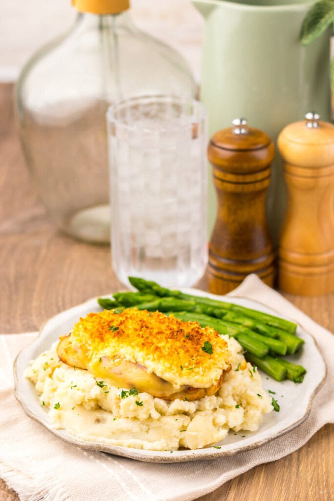 A plate of baked chicken with a crispy topping served over mashed potatoes, with steamed asparagus on the side. A glass of water and salt and pepper shakers are in the background.