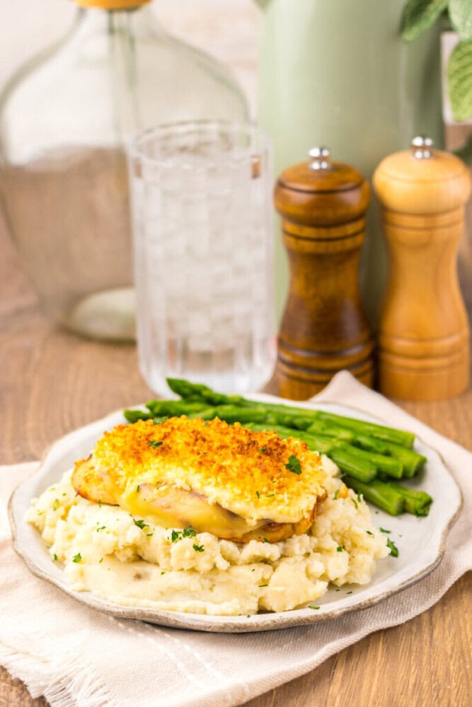 A plate of baked chicken with a crispy topping served on mashed potatoes with a side of green beans; glass of water and salt and pepper shakers in the background.