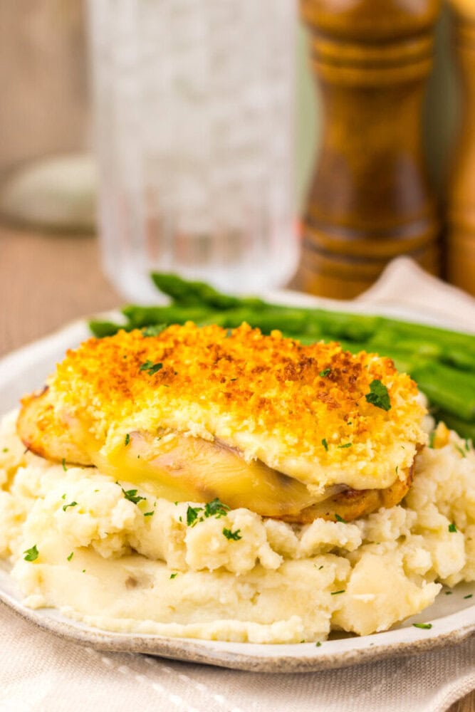 A serving of baked chicken with a golden breadcrumb crust on mashed potatoes, garnished with parsley, with green beans on the side.