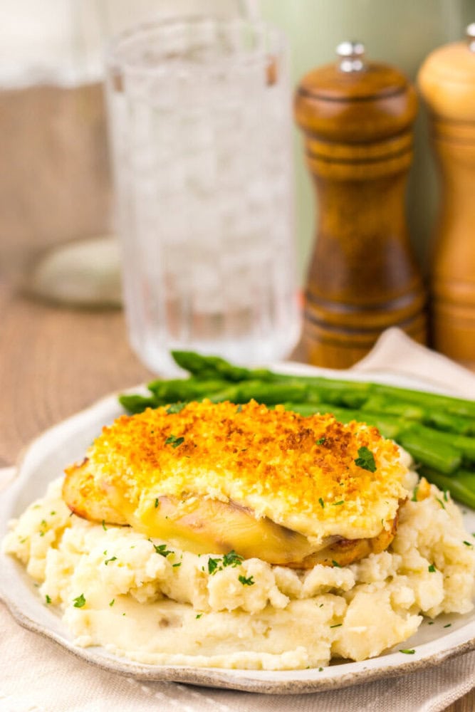 A plate of baked chicken with a crispy topping served on mashed potatoes, accompanied by asparagus spears. A glass of ice water and pepper and salt shakers are in the background.