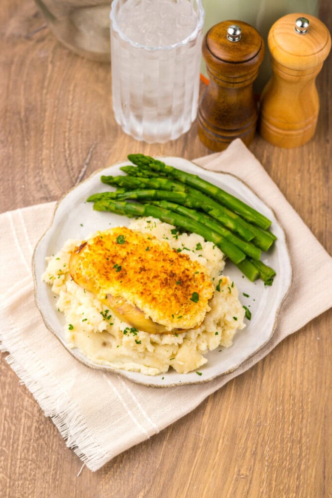 A plate of baked chicken breast on mashed potatoes with steamed asparagus, beside a glass of water and salt and pepper shakers, on a wooden table.