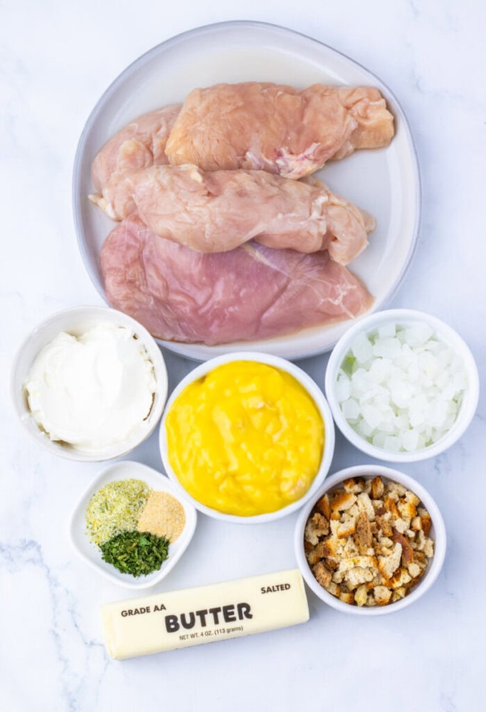 A plate of raw chicken breasts with small bowls of cream cheese, condensed soup, chopped onions, dried herbs, breadcrumbs, and a stick of salted butter on a marble surface.