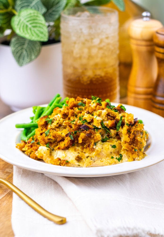 A plate of casserole with a golden breadcrumb topping and green beans, placed on a white napkin with a gold fork, a drink, and a potted plant in the background.