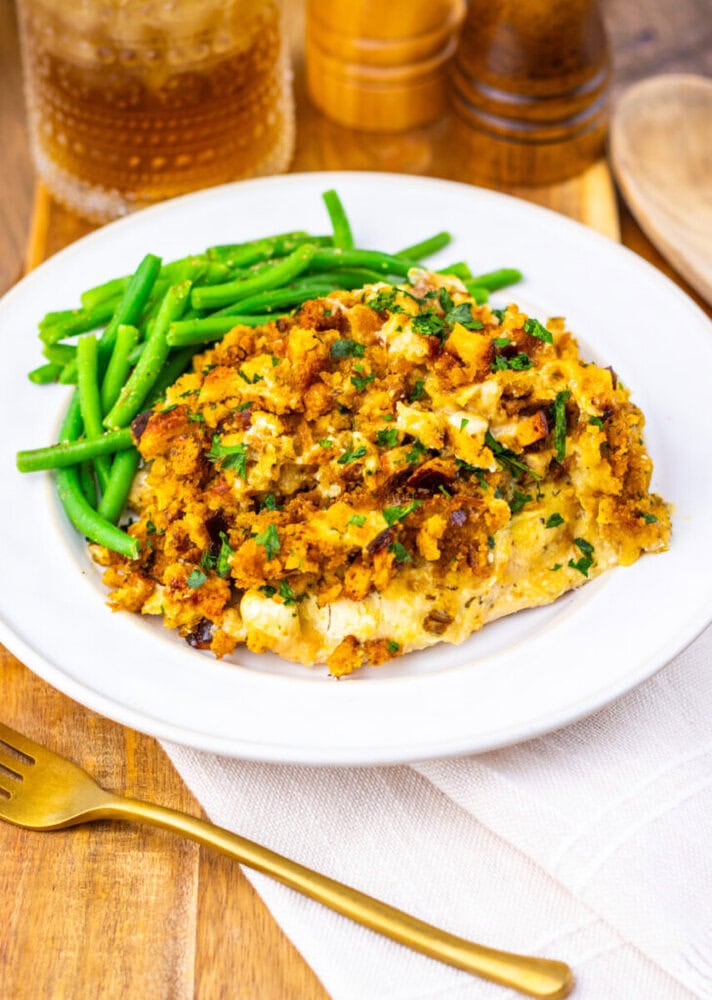 A white plate with green beans and a serving of casserole topped with golden breadcrumbs, set on a wooden table with a gold fork and a glass of iced drink.