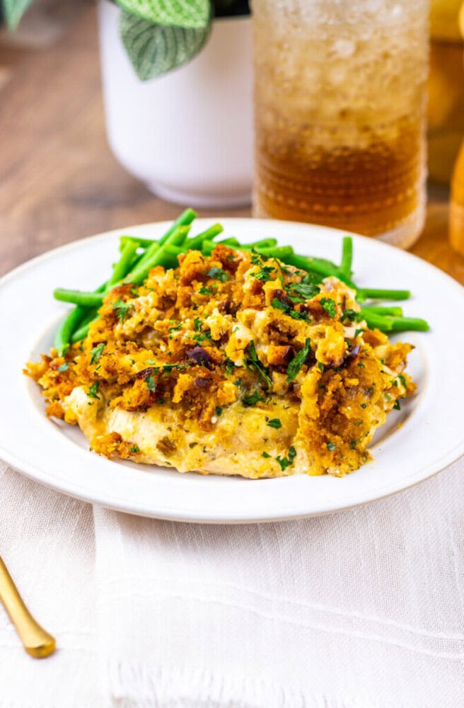 A plate of green beans and a serving of cheesy chicken casserole with a golden breadcrumb topping on a white plate, placed on a white napkin.