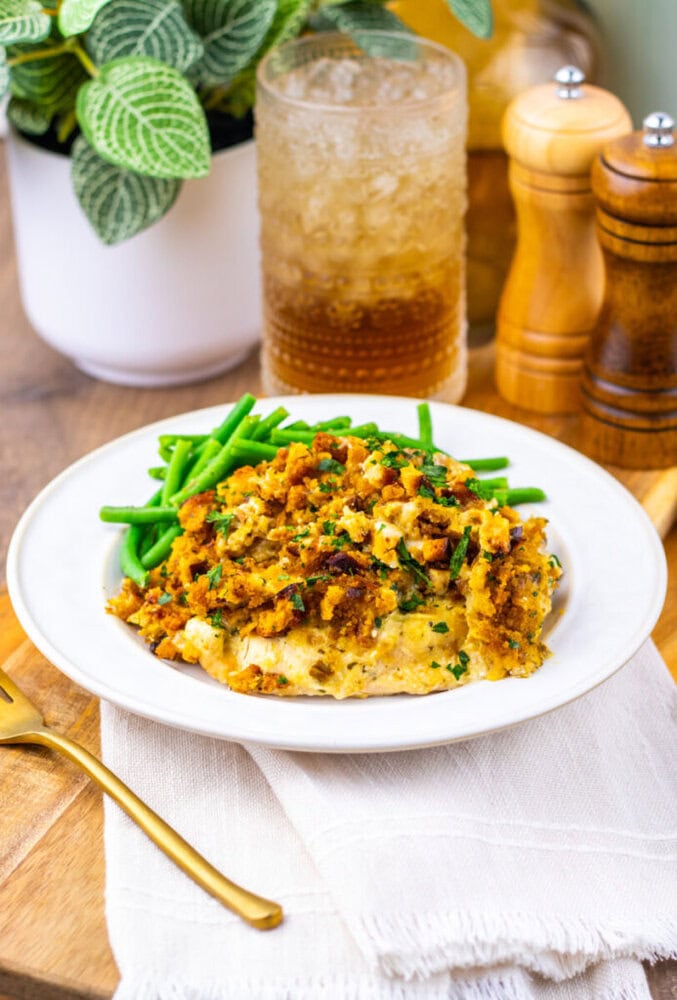 A plate of green beans and a casserole topped with breadcrumbs is served on a white dish, with a fork, glass of iced drink, salt and pepper shakers, and a potted plant nearby.