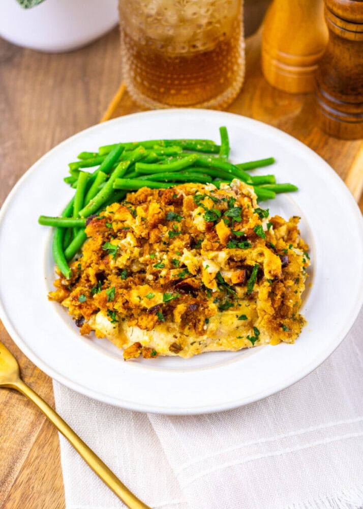 A white plate with green beans and a serving of baked fish topped with a golden breadcrumb and herb crust, placed on a wooden table with gold cutlery nearby.