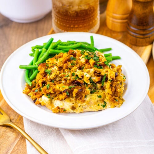 A white plate with baked casserole topped with breadcrumbs and herbs, served with green beans on the side. A fork, wooden spoon, and condiments are nearby on a wooden table.
