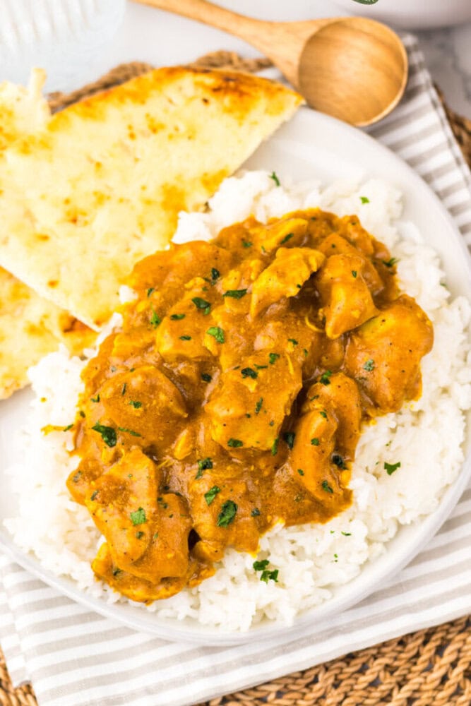 A plate of white rice topped with chicken curry garnished with herbs, served with two pieces of naan bread on the side.