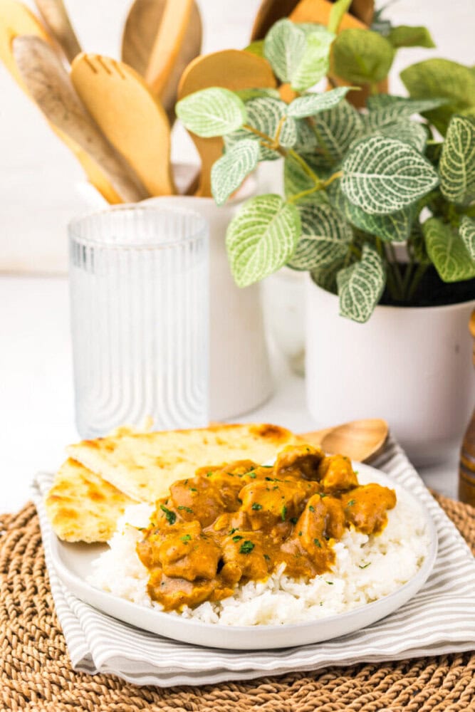 A plate of white rice topped with curry, served with naan bread and a wooden spoon, with a glass, wooden utensils, and potted plants in the background.