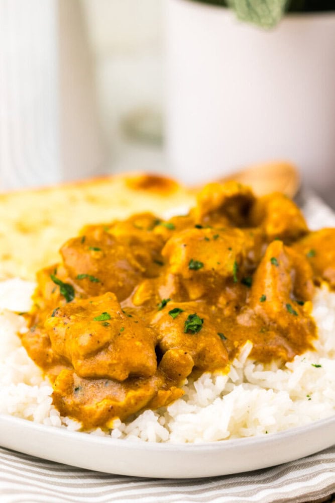 A plate of white rice topped with creamy orange curry chicken, garnished with chopped herbs, and a piece of flatbread in the background.