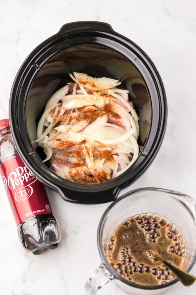 Slow cooker with raw pork, sliced onions, and spices inside, next to a bottle of Dr Pepper and a measuring cup filled with a foamy brown liquid.