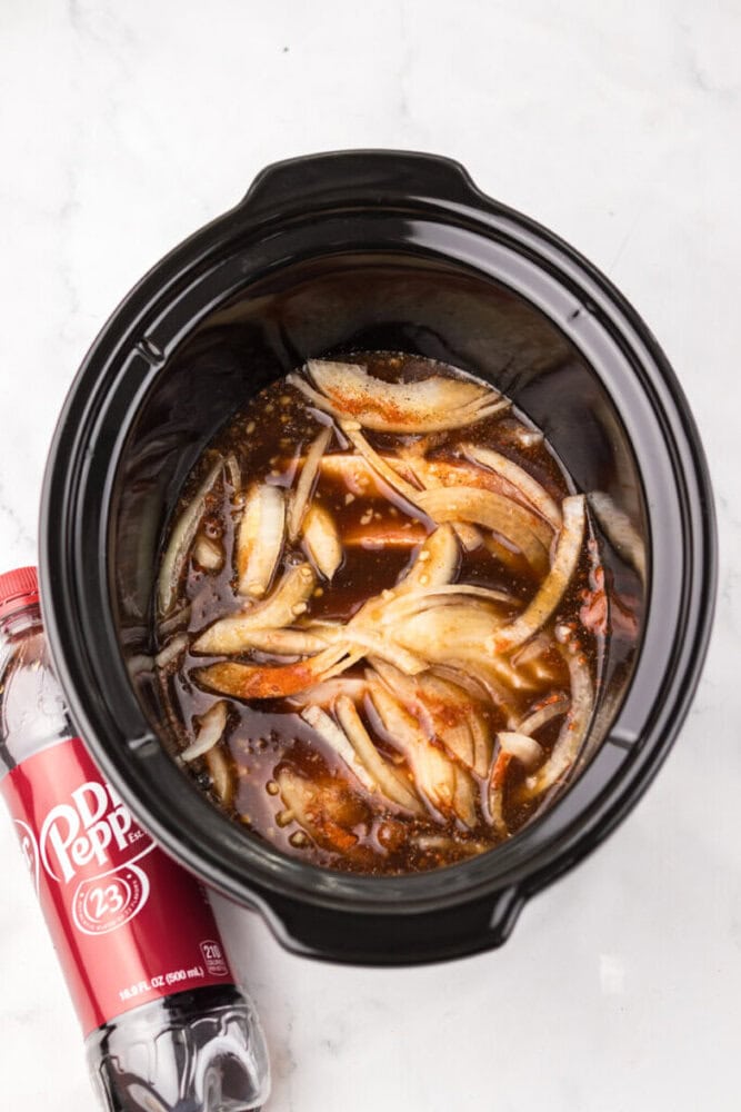 A slow cooker filled with sliced onions and a brown sauce mixture, next to an unopened bottle of Dr Pepper on a white surface.