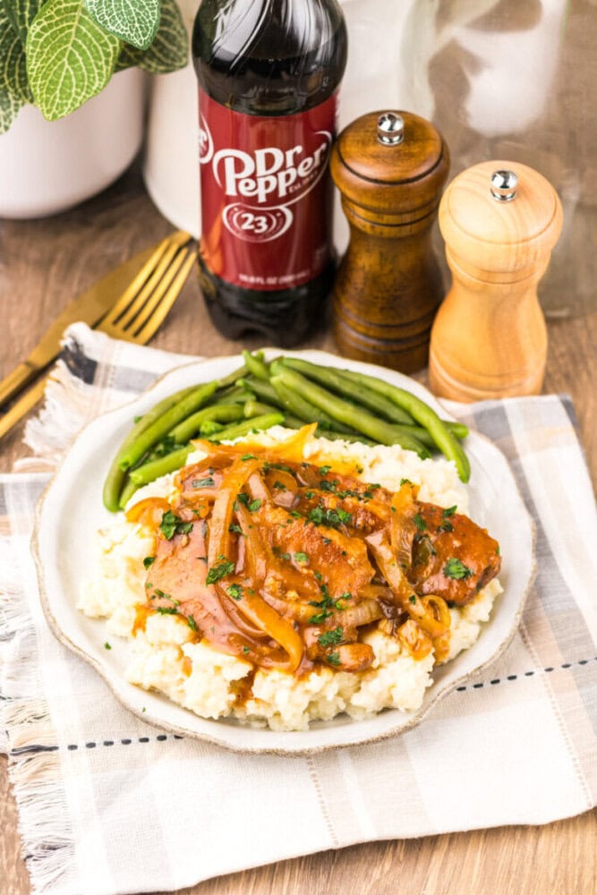 A plate of mashed potatoes topped with pork chops and onions, served with green beans. A bottle of Dr. Pepper and salt and pepper shakers are in the background.