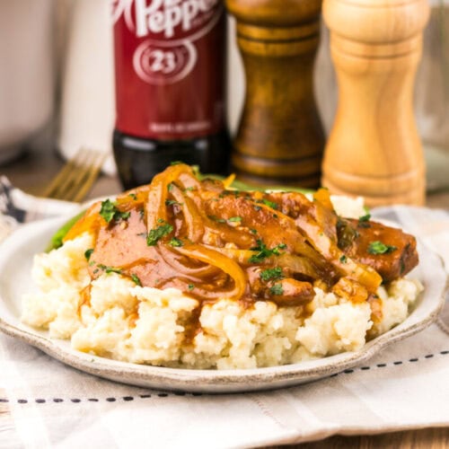 A plate of mashed potatoes topped with sliced meat, onions, and gravy sits on a cloth napkin; salt, pepper grinders, and a Dr Pepper bottle are in the background.