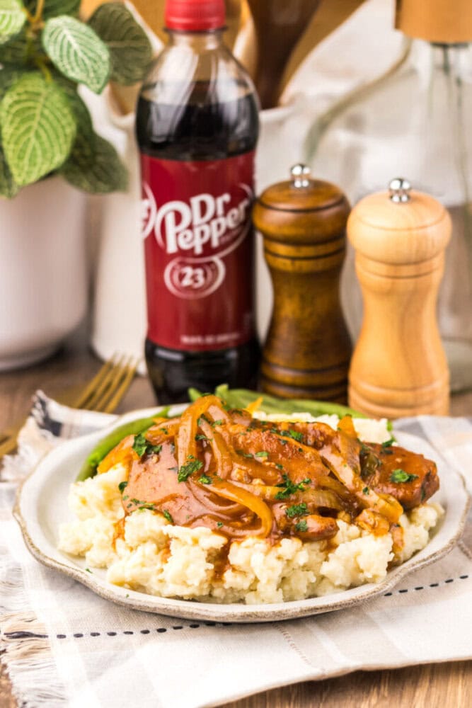 A plate of mashed potatoes topped with meat and saut&eacute;ed onions, with a bottle of Dr Pepper and salt and pepper shakers in the background.