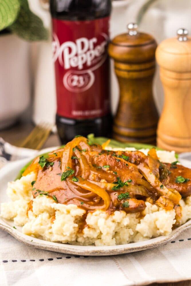 A plate of mashed potatoes topped with meat, saut&eacute;ed onions, and sauce, with a bottle of Dr Pepper and salt and pepper shakers in the background.